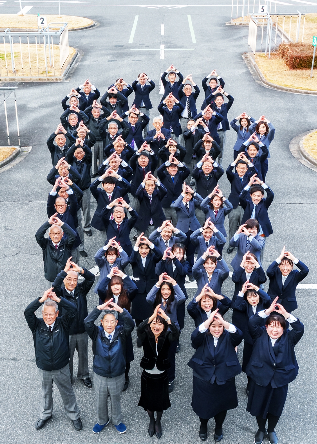 自動車学校の職員が屋外で並び、手でポーズをしている集合写真